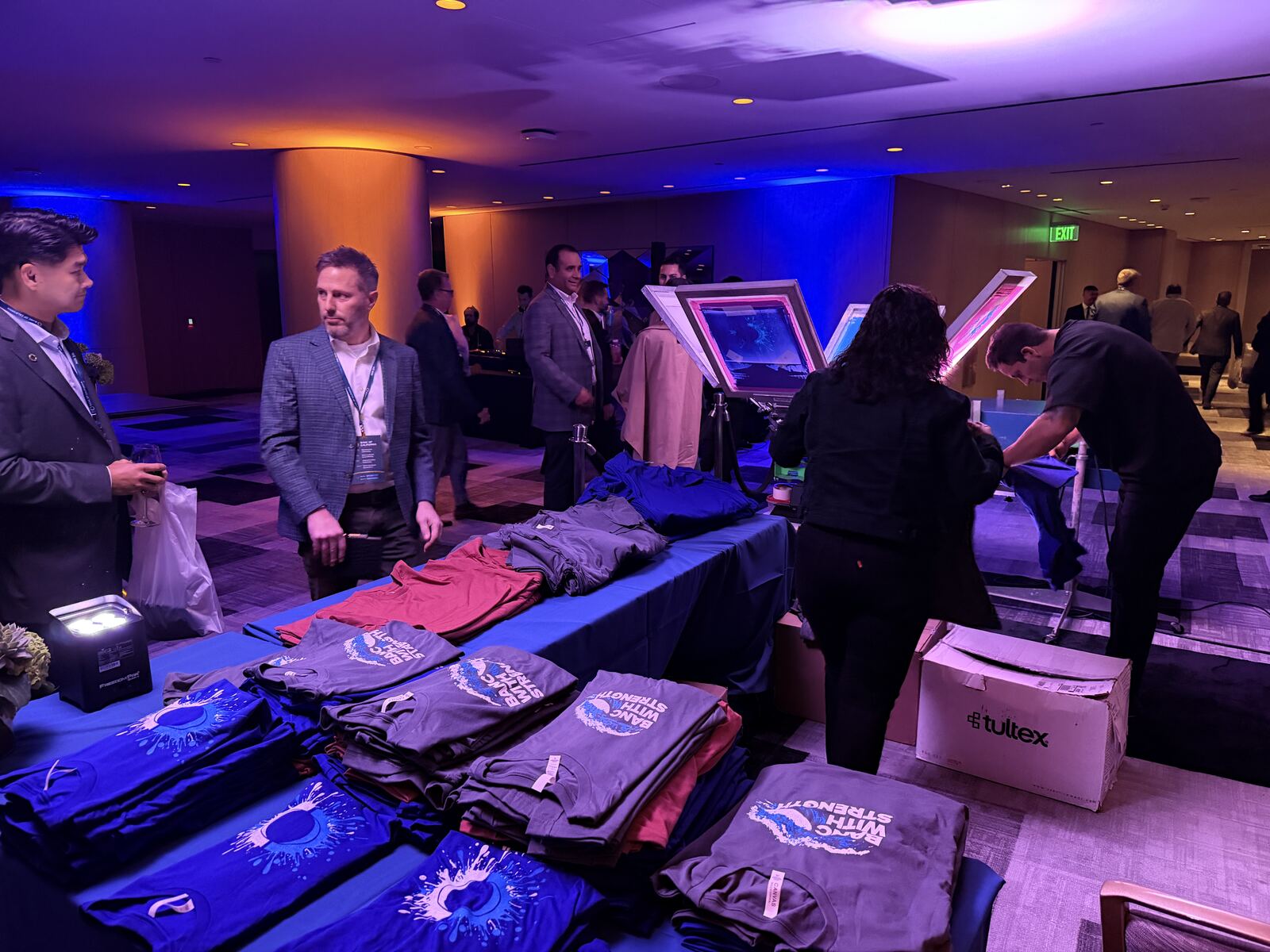Indoor event venue with attendees walking past merchandise tables displaying branded apparel